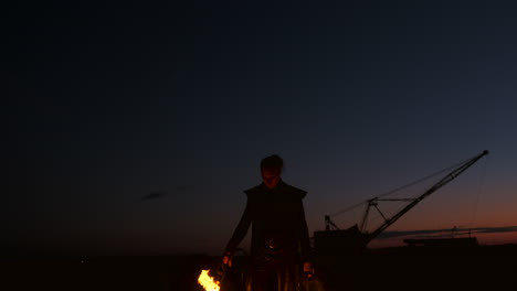 Ein-Mann-In-Einem-Regenmantel-Mit-Zwei-Flammenwerfern-Lässt-Eine-Feurige-Flamme-Aus,-Die-Bei-Sonnenuntergang-Auf-Dem-Sand-Steht